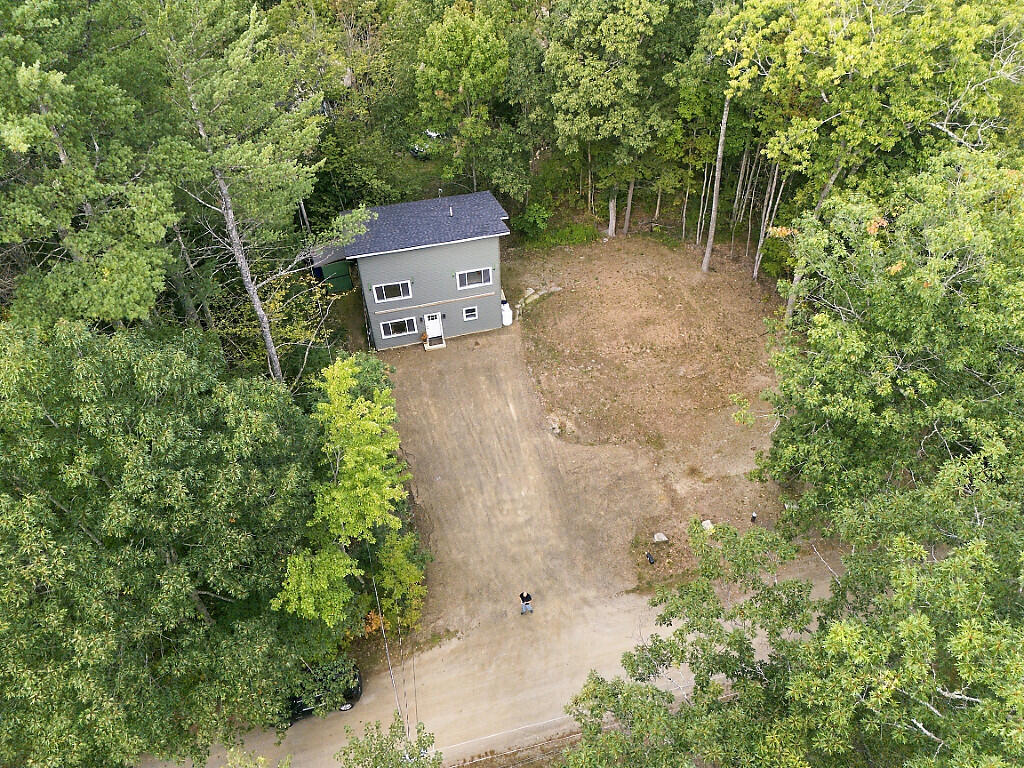 Aerial view of a gray two-story house surrounded by trees, with a dirt driveway and cleared yard.