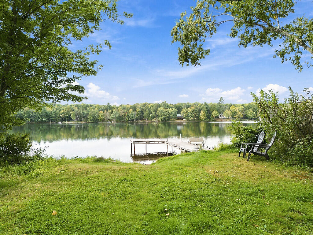 Lakeside yard with a wooden dock, two chairs, and calm water backed by trees.