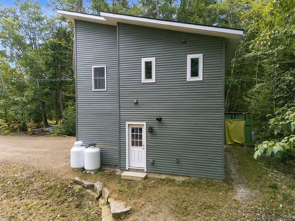 Gray-sided home with a steep shed-style roof, side entry door, and propane tanks among trees.