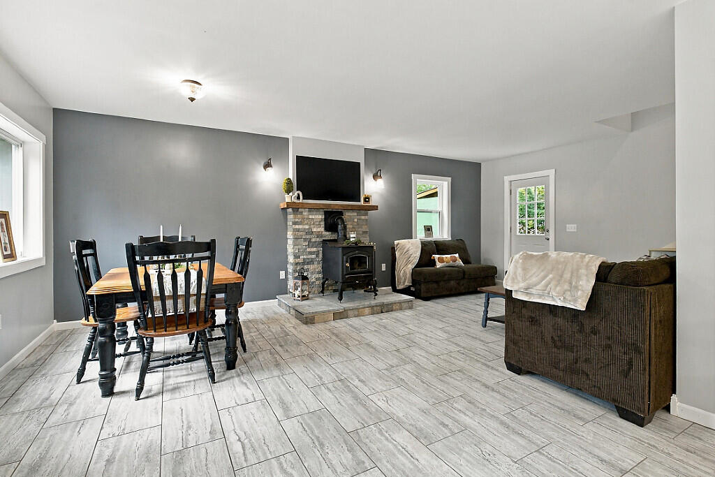 Open-plan living and dining room with a stone fireplace, wood-burning stove, and gray tile floor.