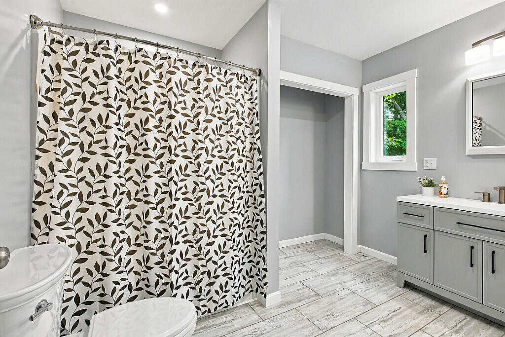 Bathroom with a leaf-patterned shower curtain, gray vanity, and gray wood-look tile floor.