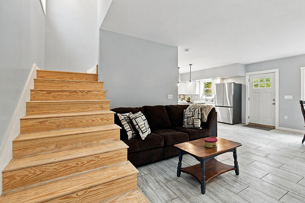 Living room with a dark sectional, wood staircase, gray flooring, and adjoining kitchen.