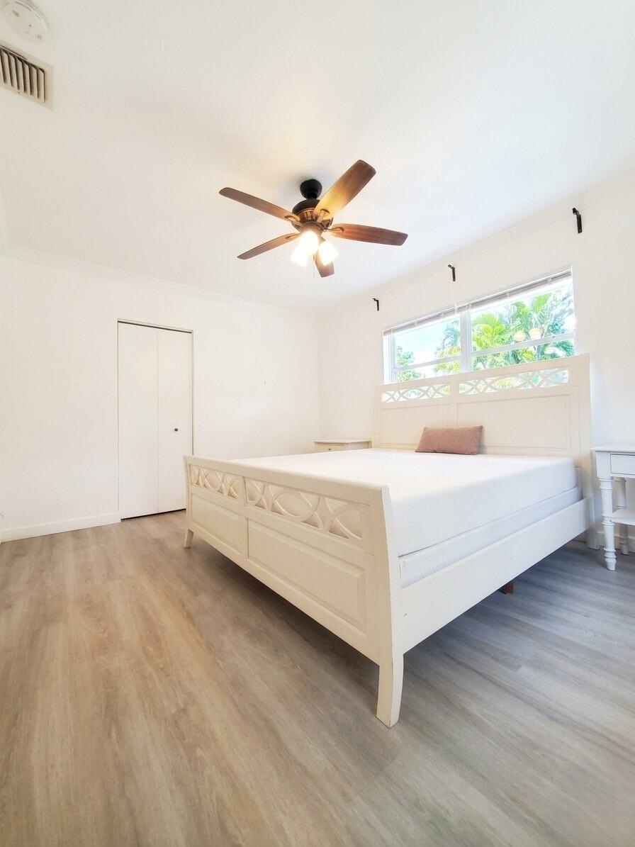 Bright bedroom with a white decorative bed, wood-look flooring, and palm trees visible through the window.