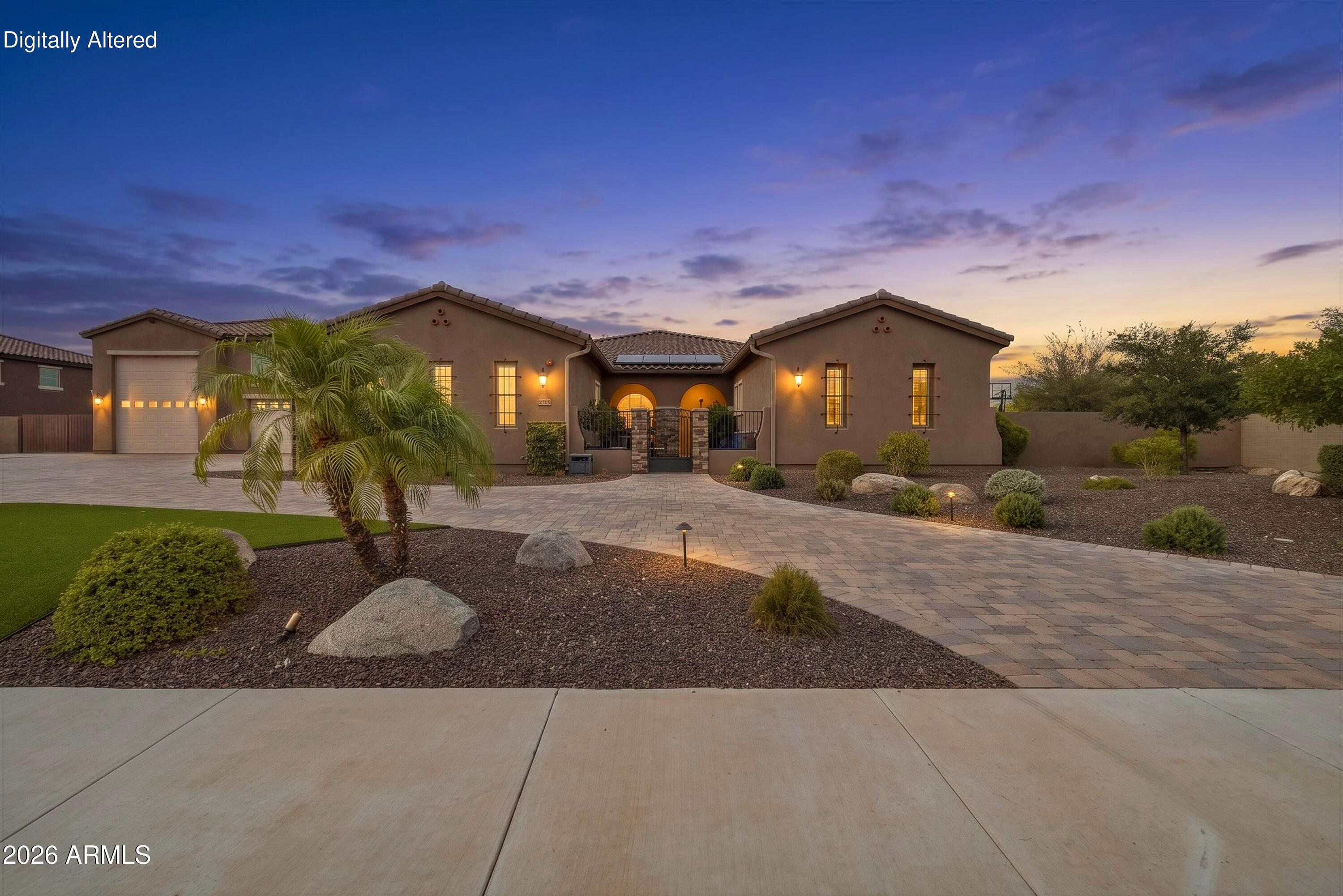Stucco home at dusk with an illuminated entry, broad paver driveway, palm trees, and desert landscaping.
