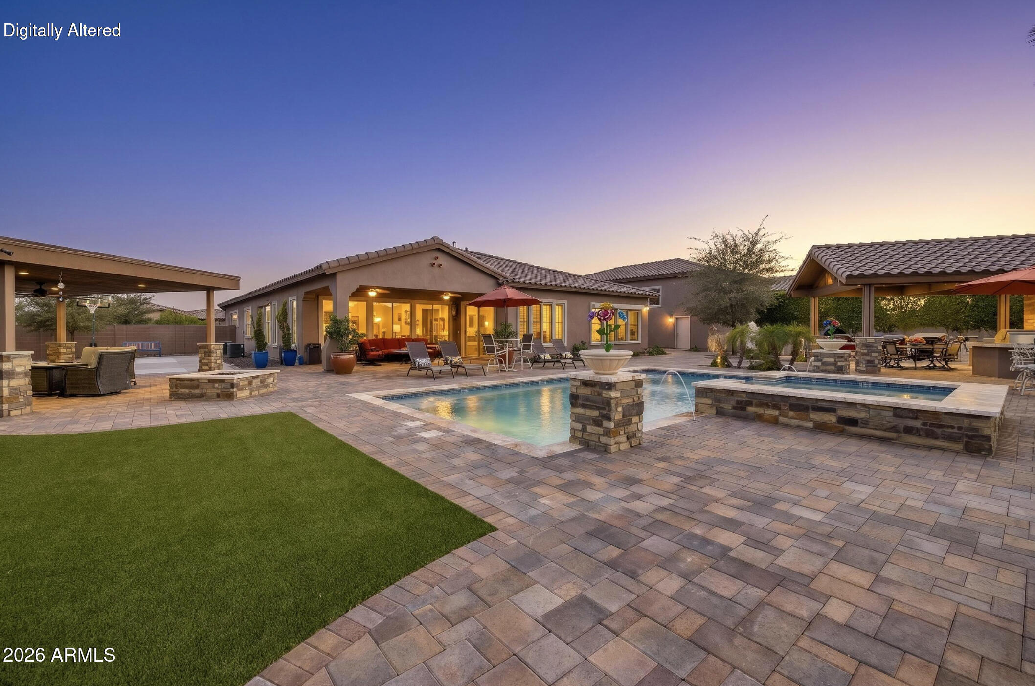 Backyard patio with illuminated swimming pool, raised spa, covered seating areas, and a green lawn at dusk.