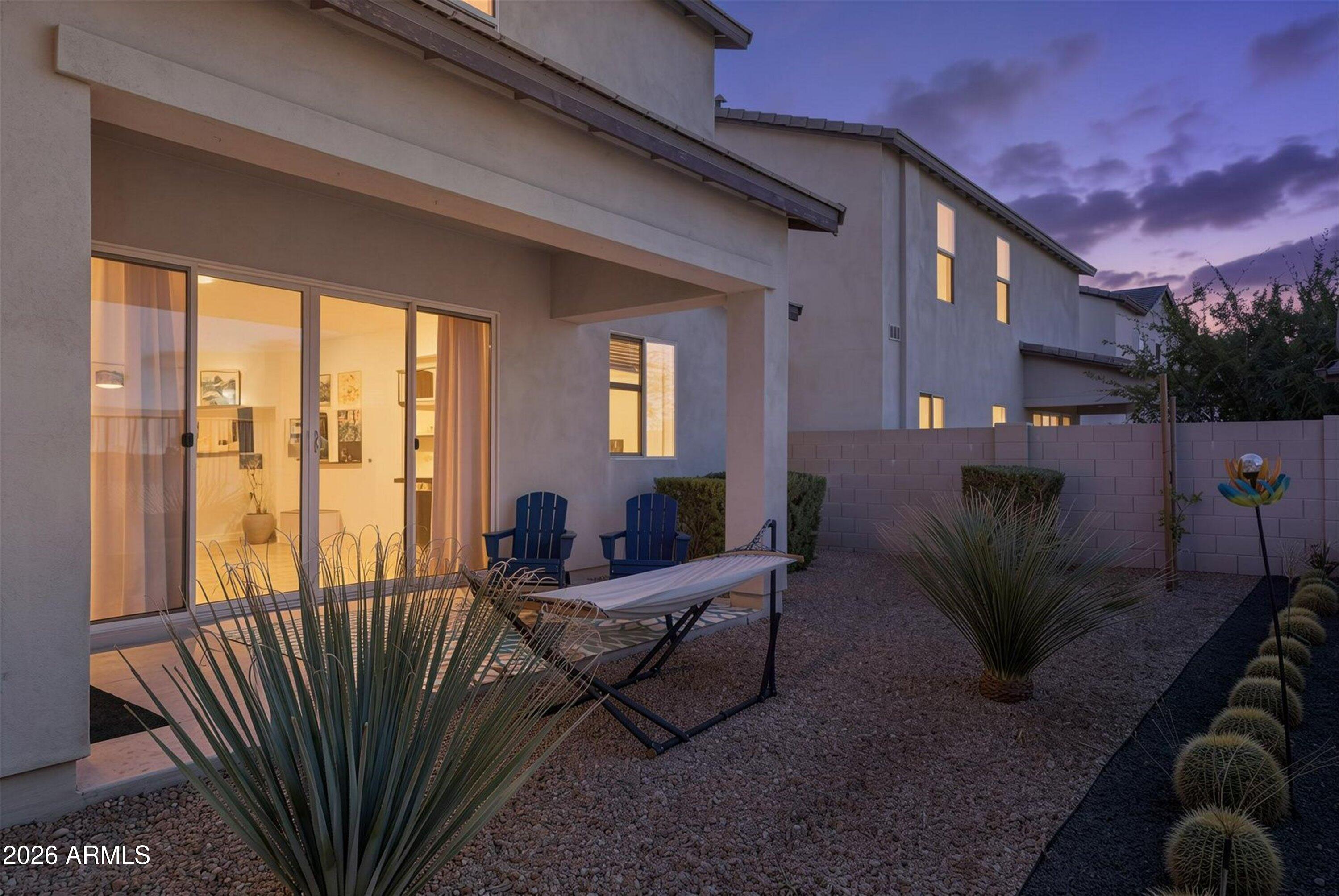 Modern home’s gravel backyard at dusk, with a hammock, blue patio chairs, and desert landscaping.