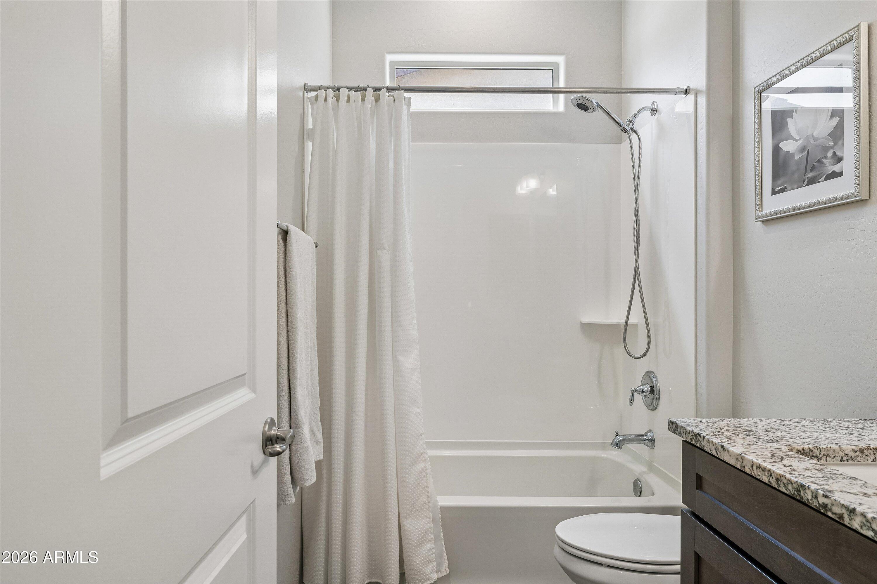 Bathroom with a white tub and shower, narrow window, toilet, and dark vanity with granite countertop.