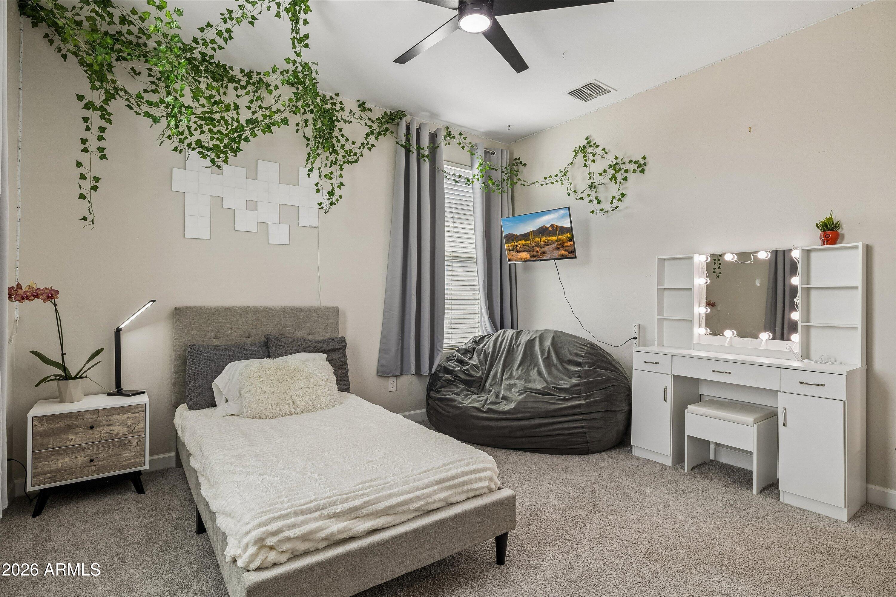 Furnished bedroom with a gray bed, beanbag chair, illuminated vanity mirror, and trailing greenery.