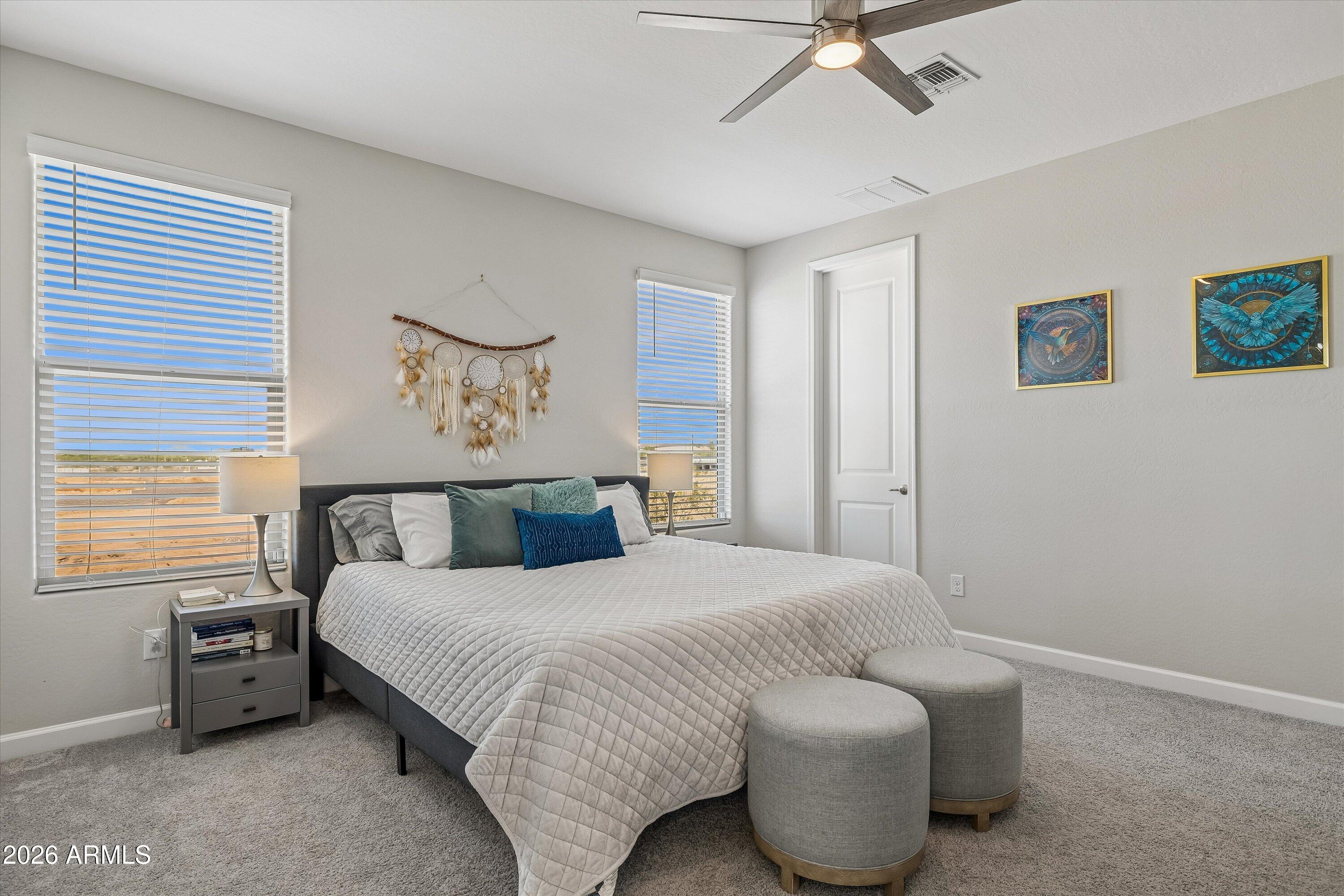 Furnished bedroom with a white-quilted bed, round stools, window blinds, and colorful wall art.