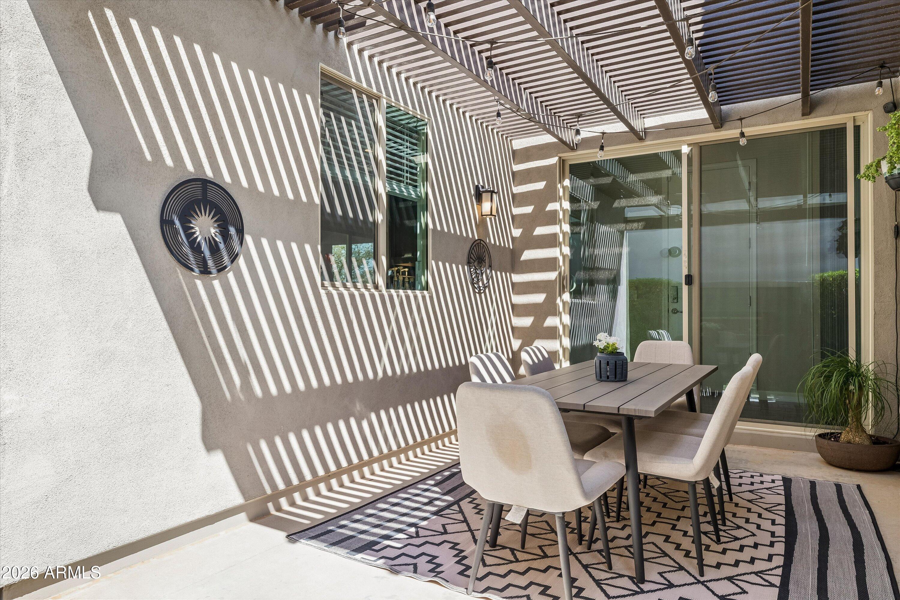 Covered patio with pergola, outdoor dining set, and patterned rug beside sliding glass doors.
