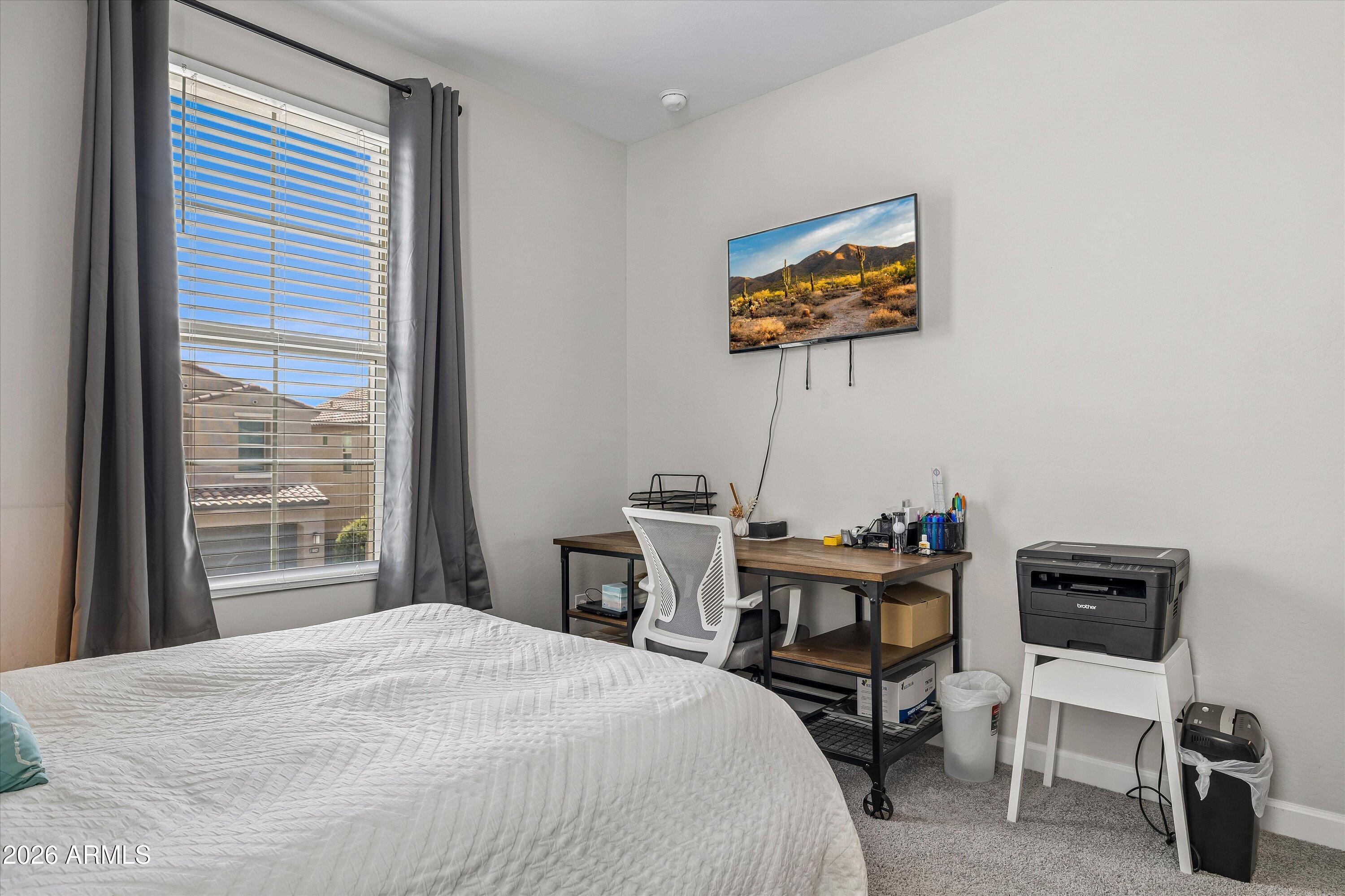 Bedroom with a desk and office chair, wall-mounted TV, printer, and window framed by gray curtains.