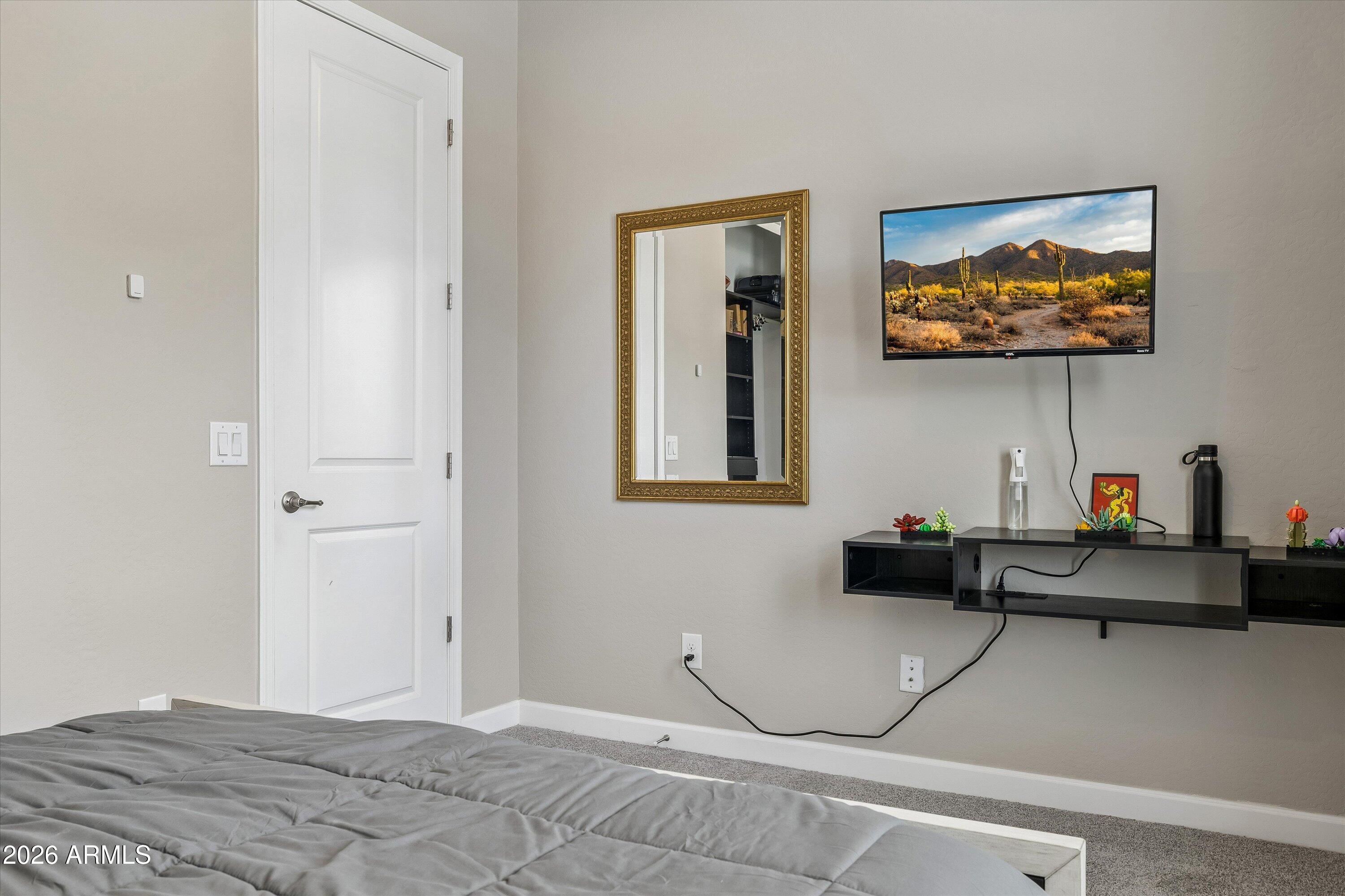 Bedroom with gray bedding, a wall-mounted television, and a gold-framed mirror.