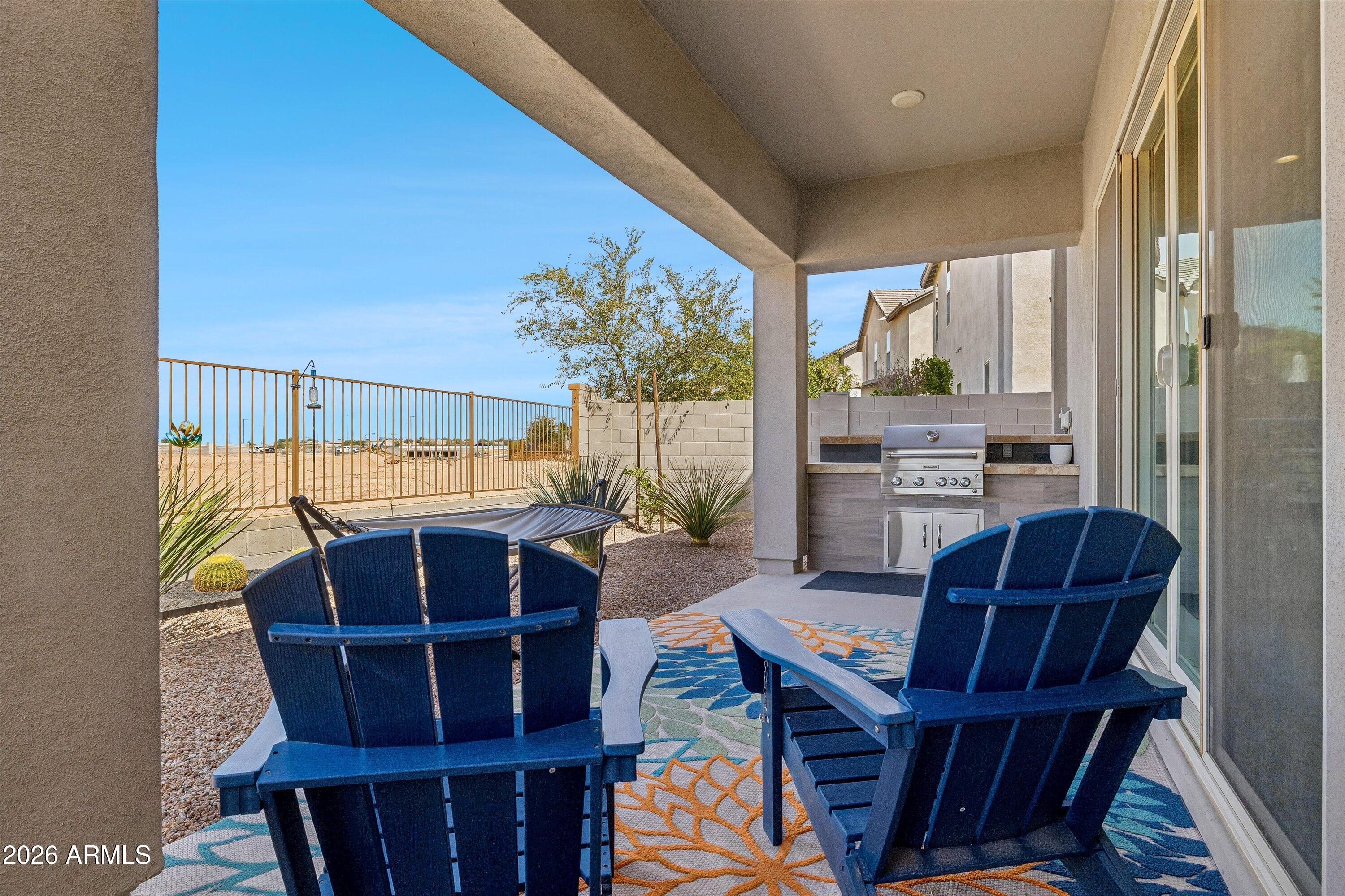 Covered patio with blue Adirondack chairs, a colorful rug, hammock, and built-in grill.