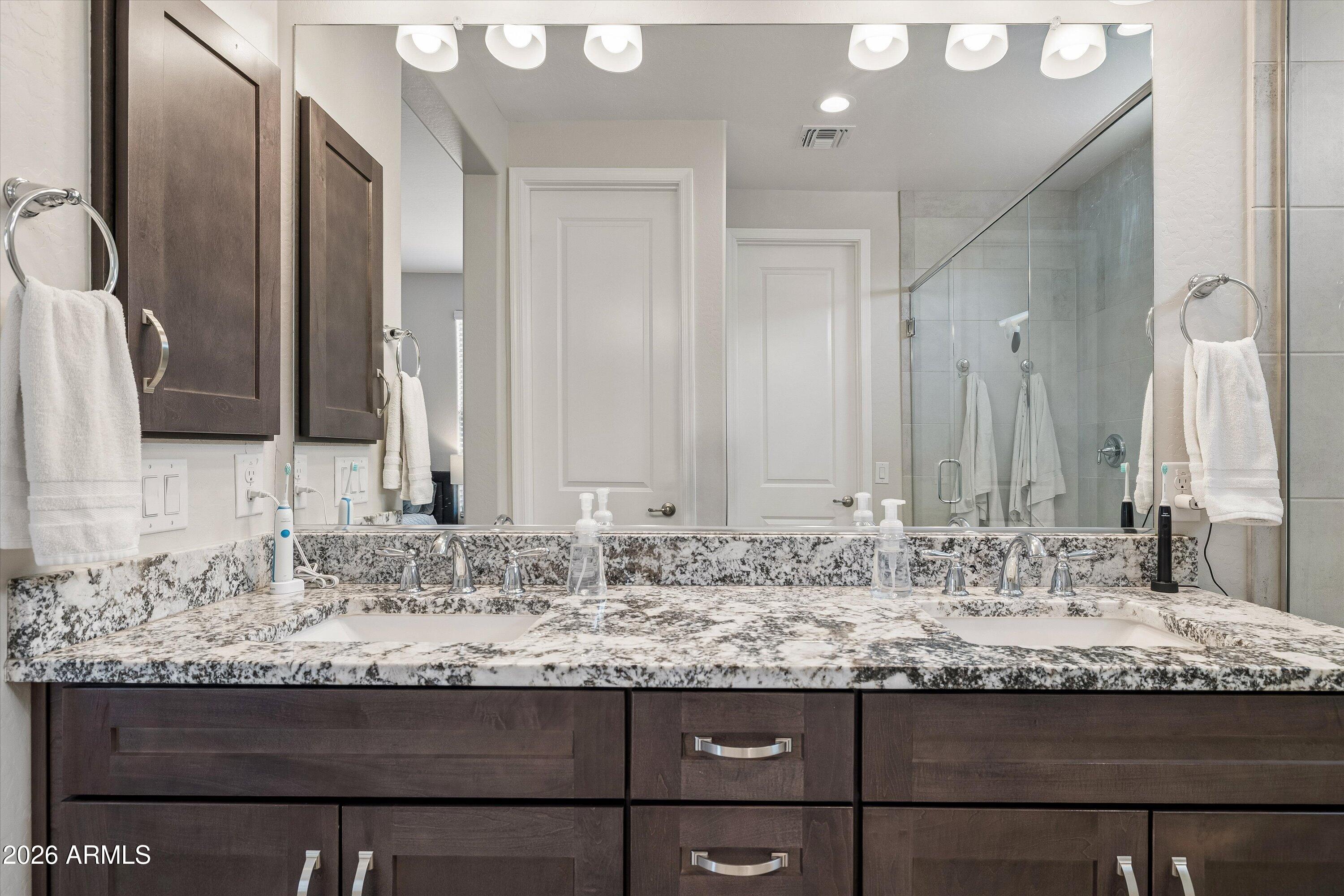 Bathroom with double sinks, dark wood vanity cabinets, speckled stone counters, and a glass-enclosed shower.