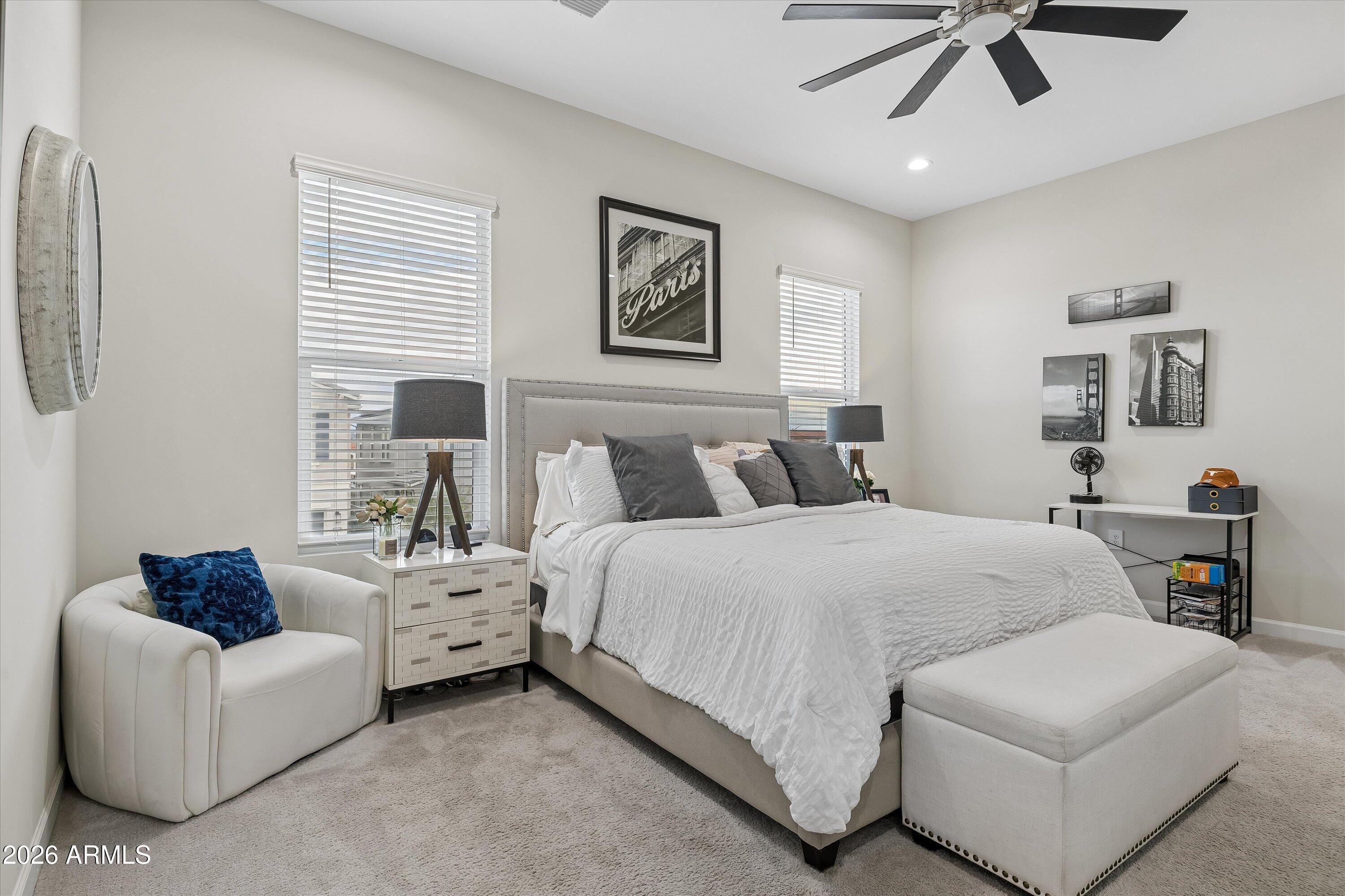 Staged bedroom with a large white bed, upholstered chair and ottoman, and dark ceiling fan.