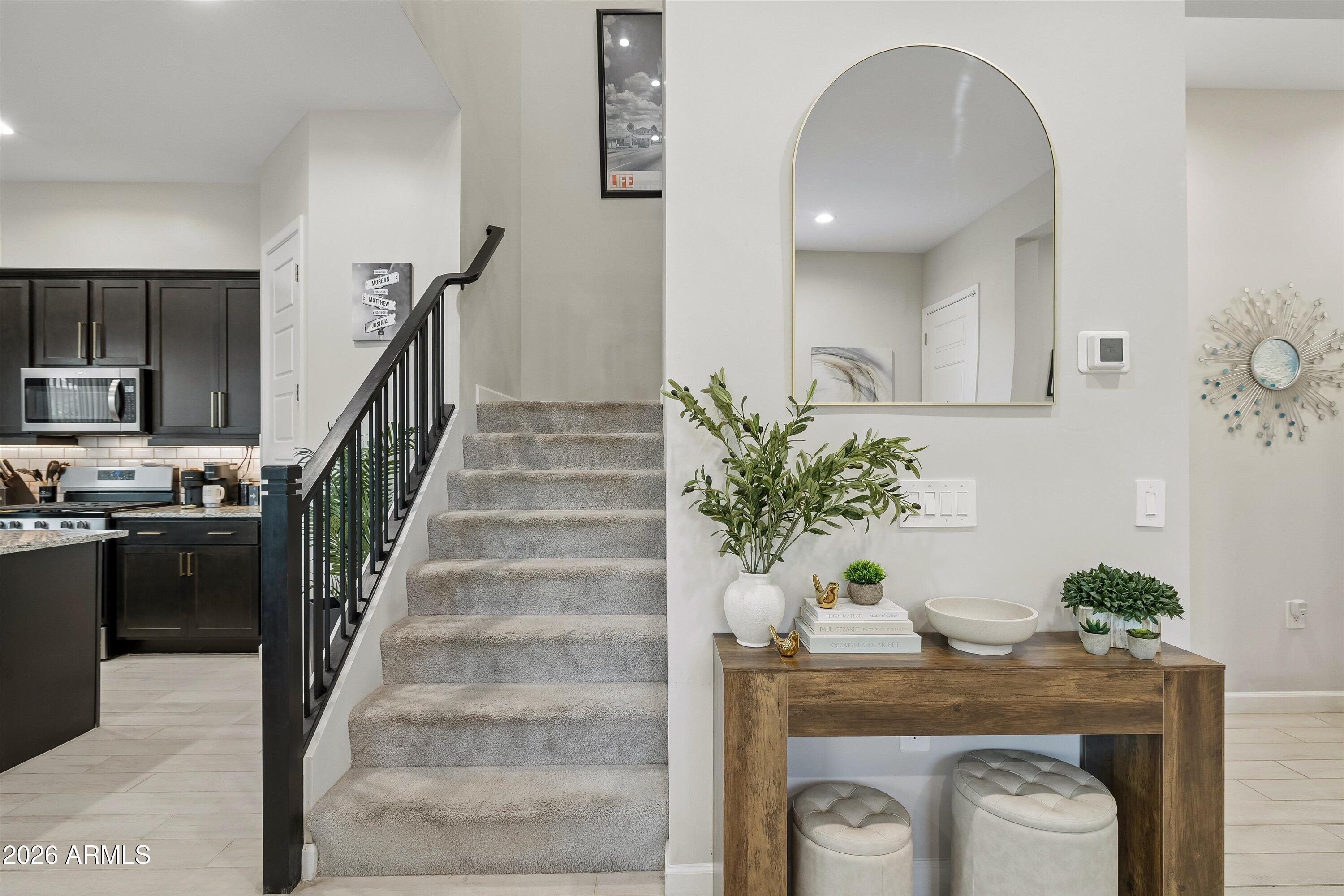 Staged entryway with carpeted stairs, a dark-cabinet kitchen, and a console table beneath an arched mirror.