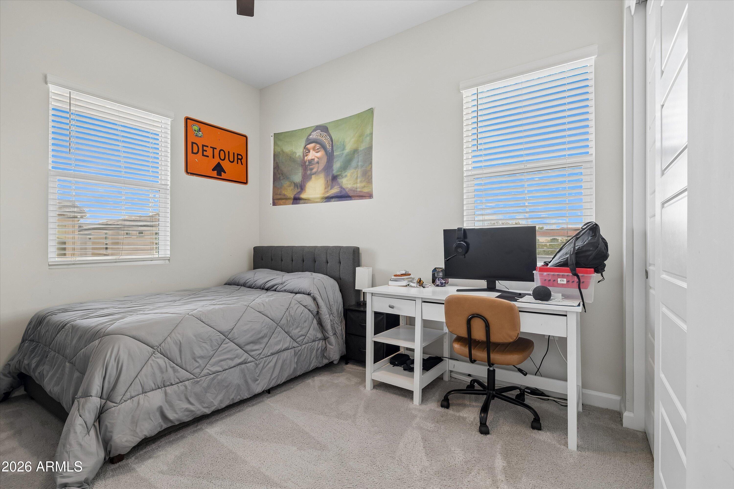 Bedroom with a gray bed, white desk and computer, two bright windows, and novelty wall signs.
