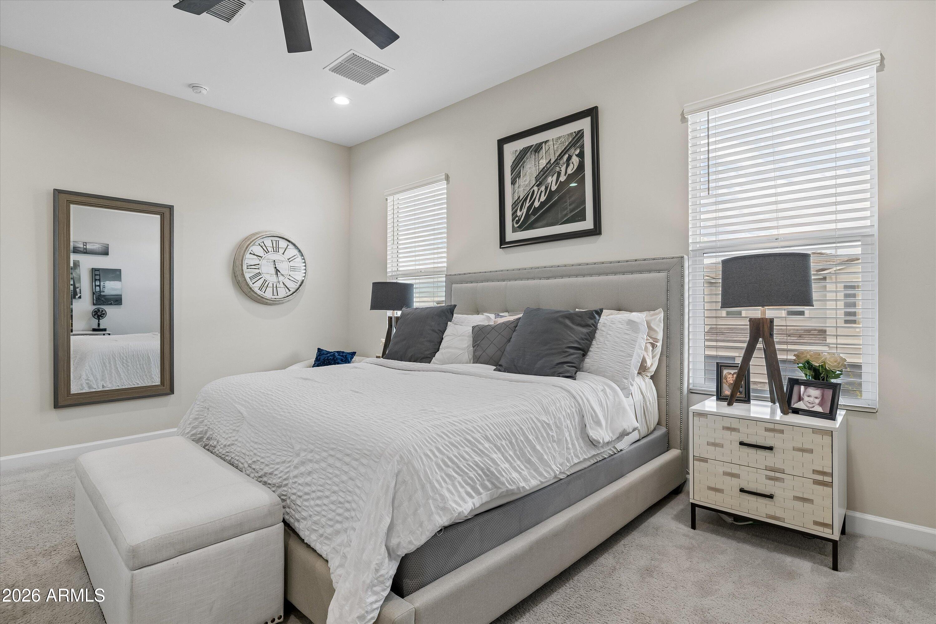 Staged bedroom with a large white bed, cushioned foot bench, and tripod lamp beside bright windows.