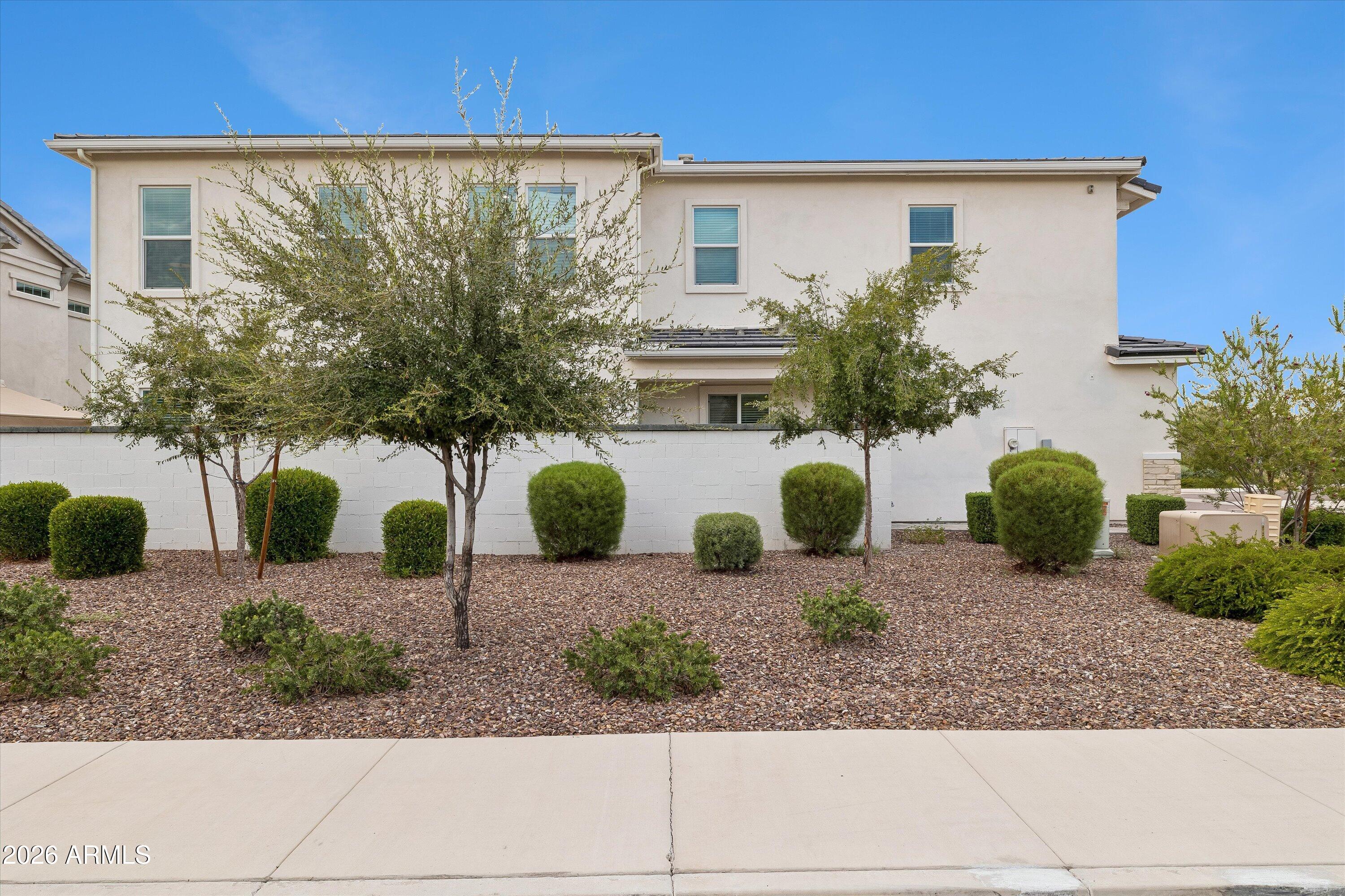Two-story stucco home behind a block wall, with gravel landscaping, shrubs, and trees.