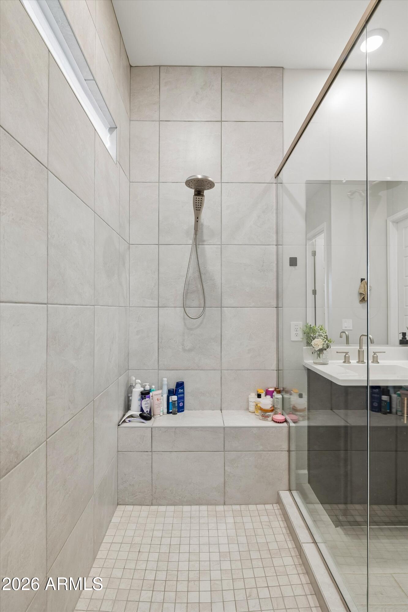 Walk-in shower with large pale tiles, glass enclosure, handheld showerhead, and built-in bench.