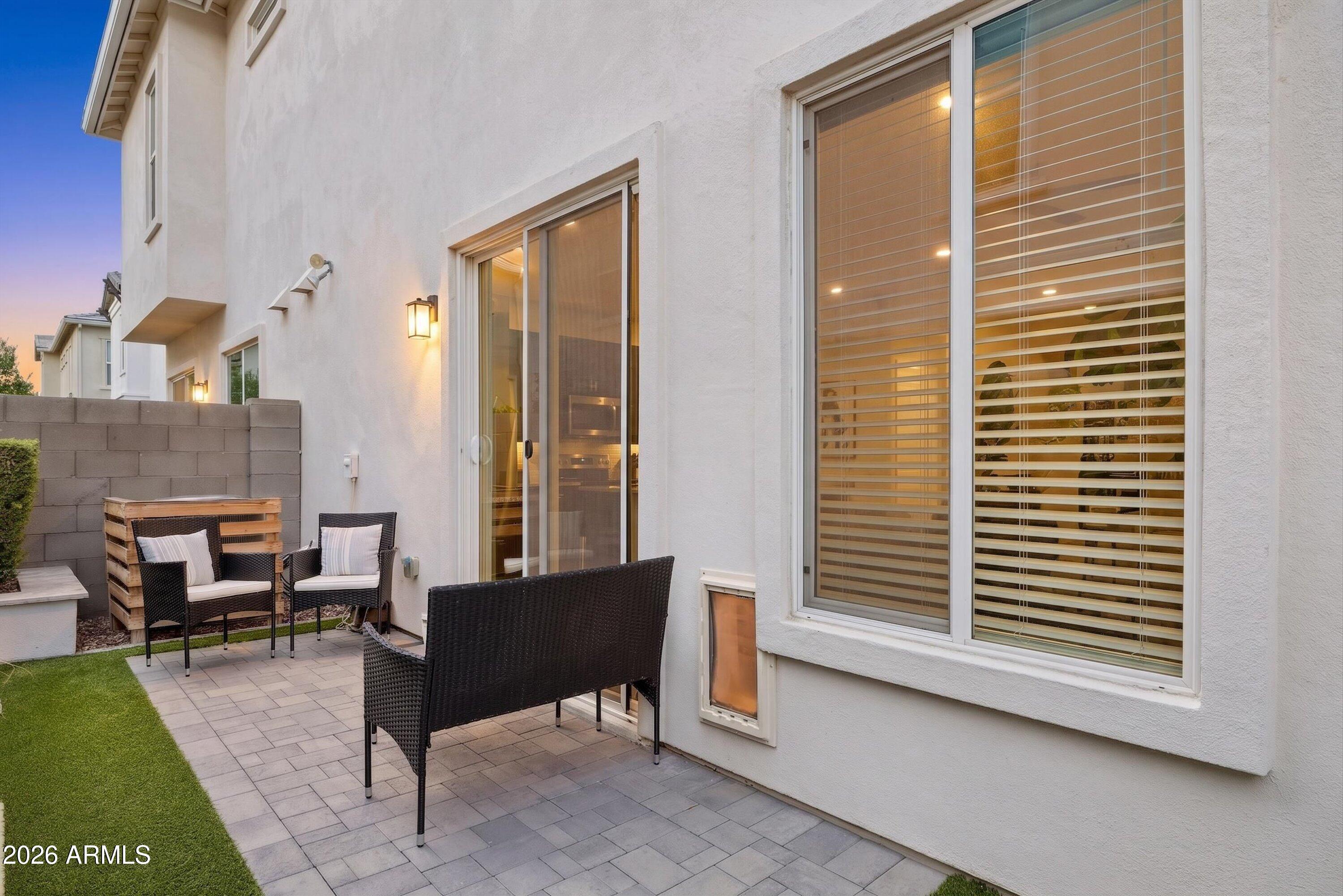 Private paved patio with woven seating and artificial turf beside a white stucco home at dusk.