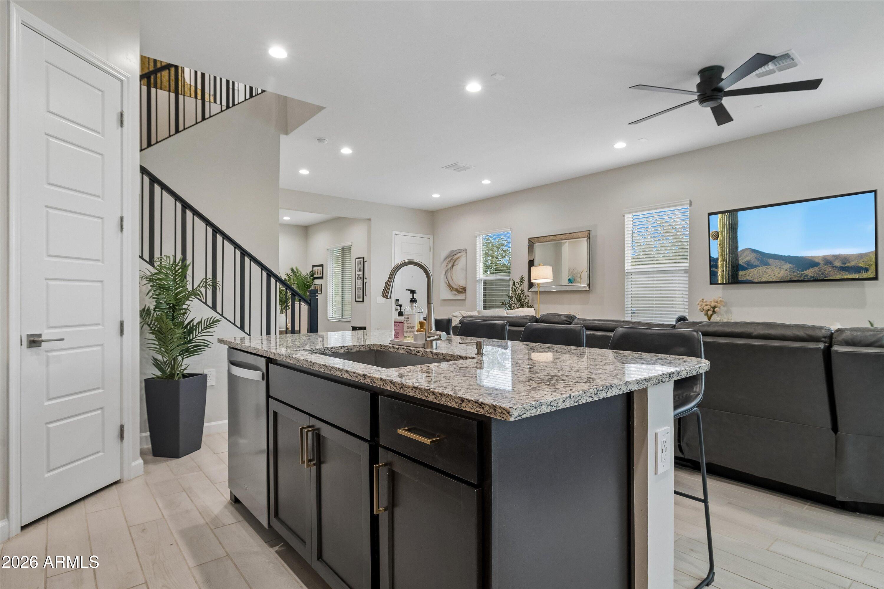 Open kitchen with a large granite island, dark cabinetry, and a black-railed staircase beside the living room.