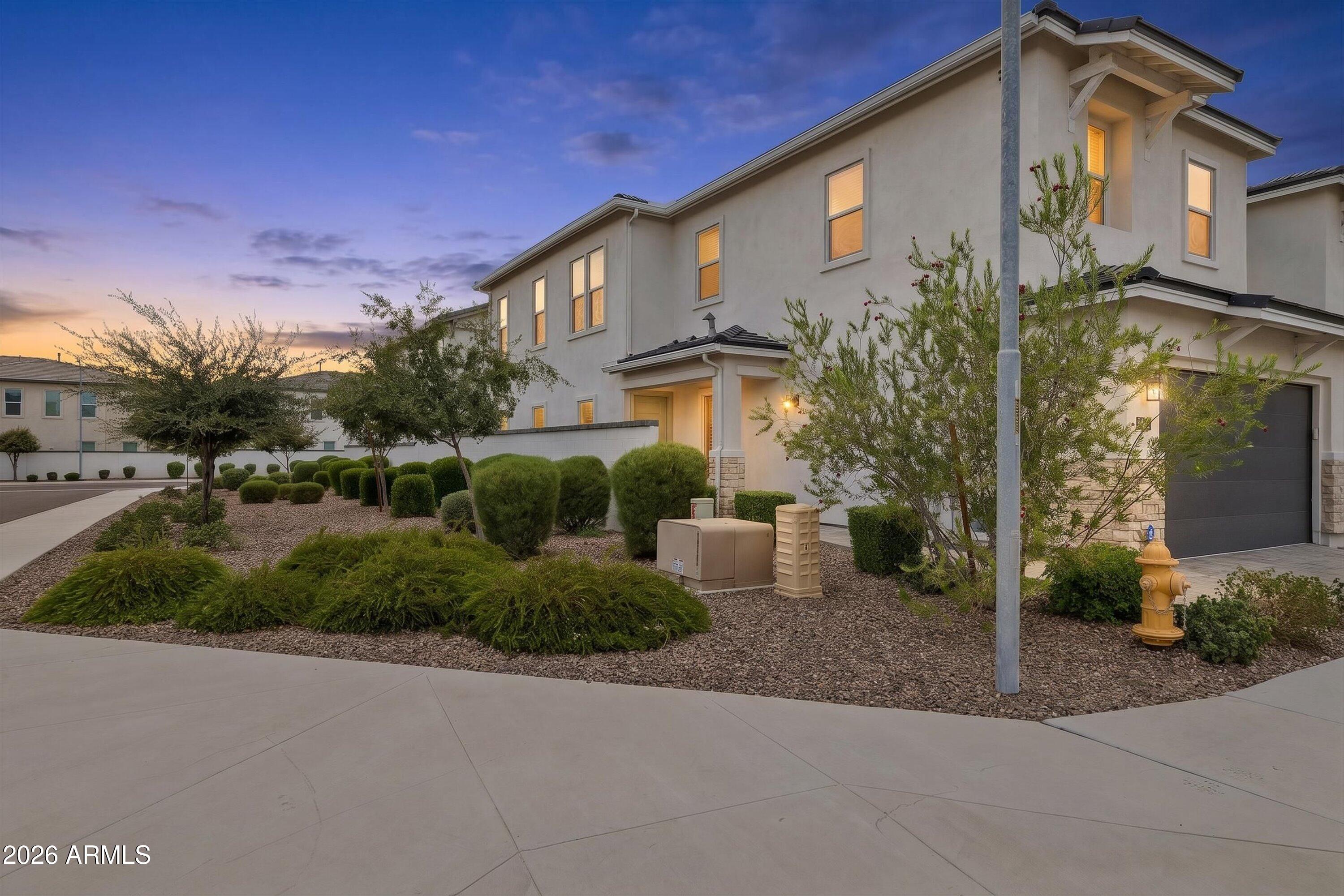 Two-story stucco home at dusk with warm-lit windows, desert landscaping, and a dark garage door.