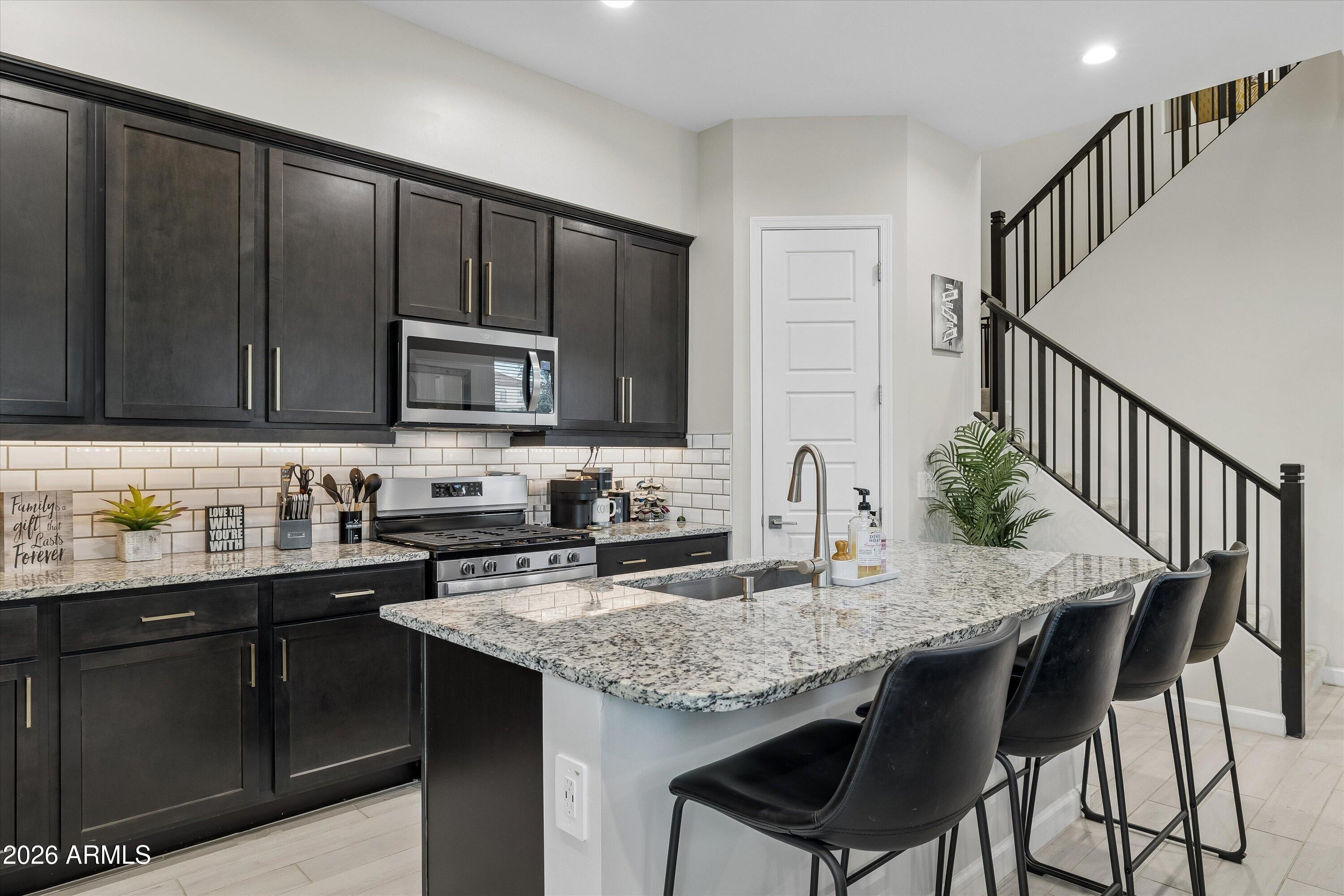 Kitchen with dark cabinets, granite island, black bar stools, and a staircase with black railings.