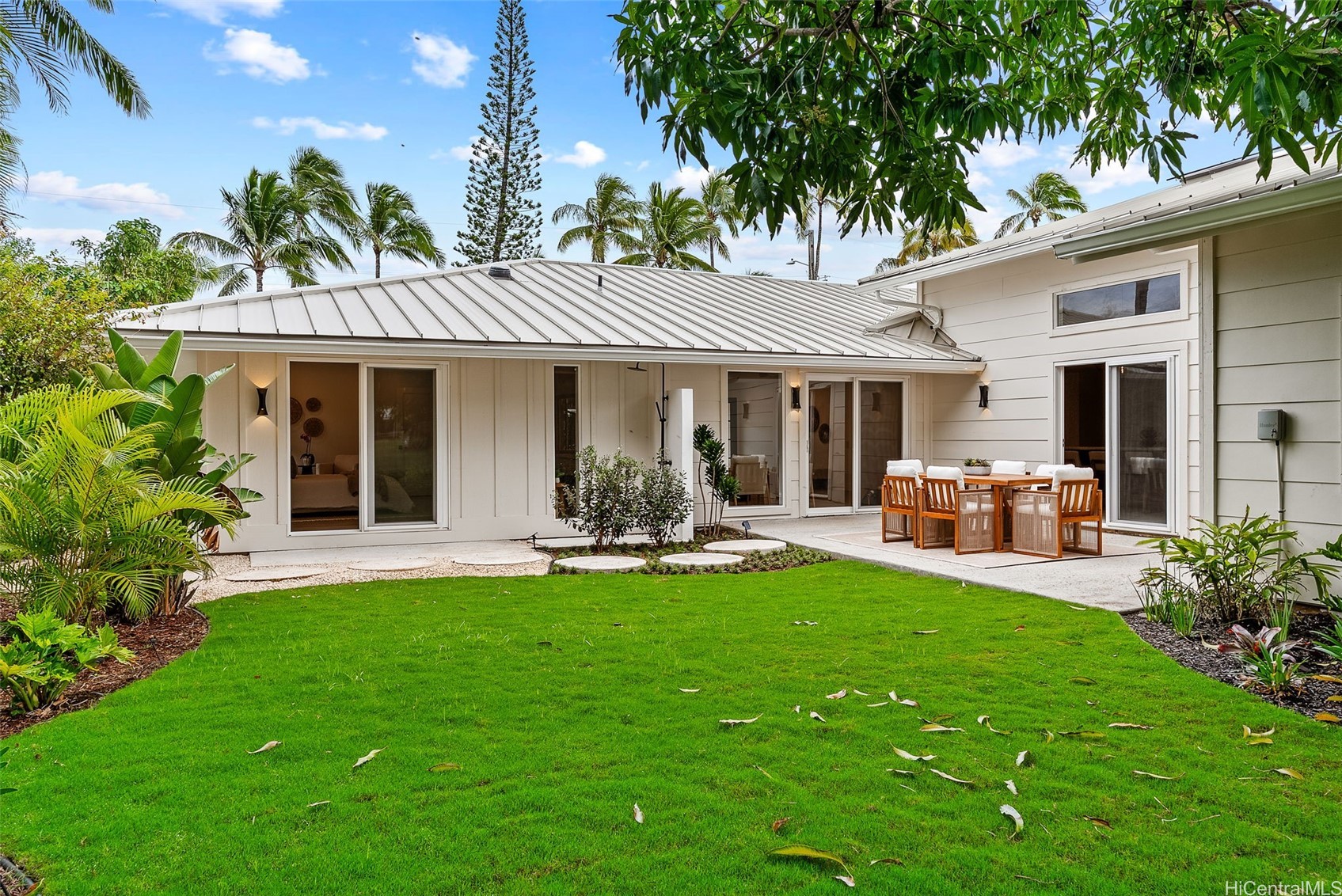 Rear of a light-colored home with a metal roof, patio dining set, and lush tropical lawn.