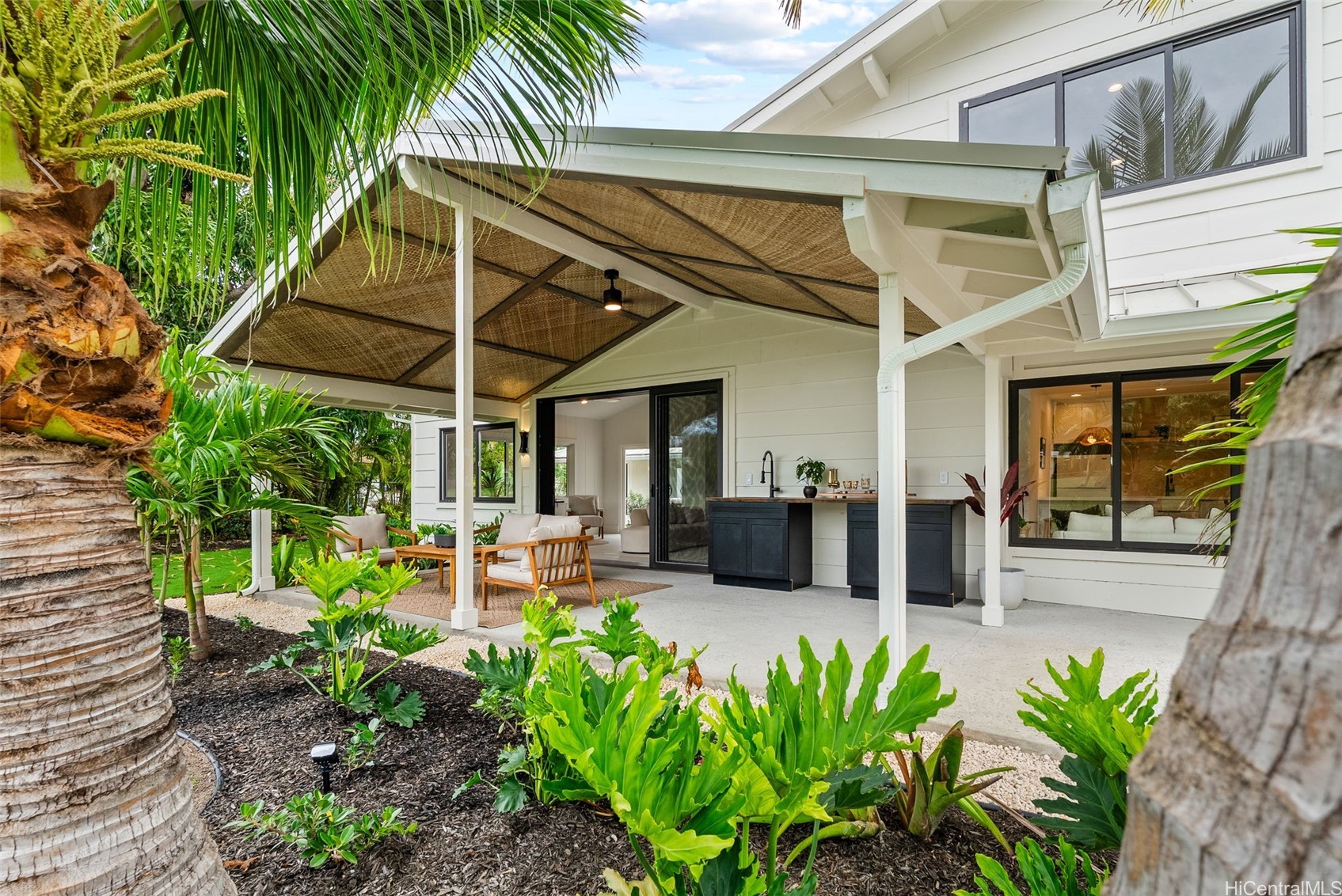 Covered lanai with woven ceiling, outdoor seating, dark kitchen counter, and lush tropical landscaping.