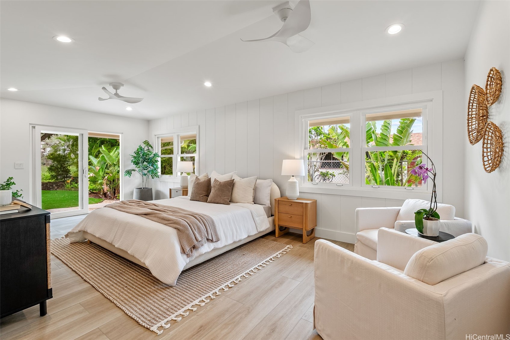 Staged bedroom with a large bed, white lounge chairs, and windows overlooking tropical greenery.