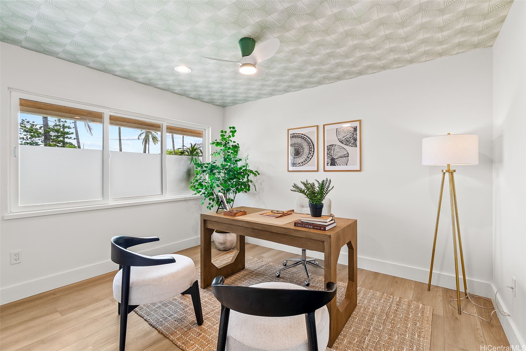 Staged home office with a wooden desk, patterned ceiling, and wide windows overlooking palm trees.
