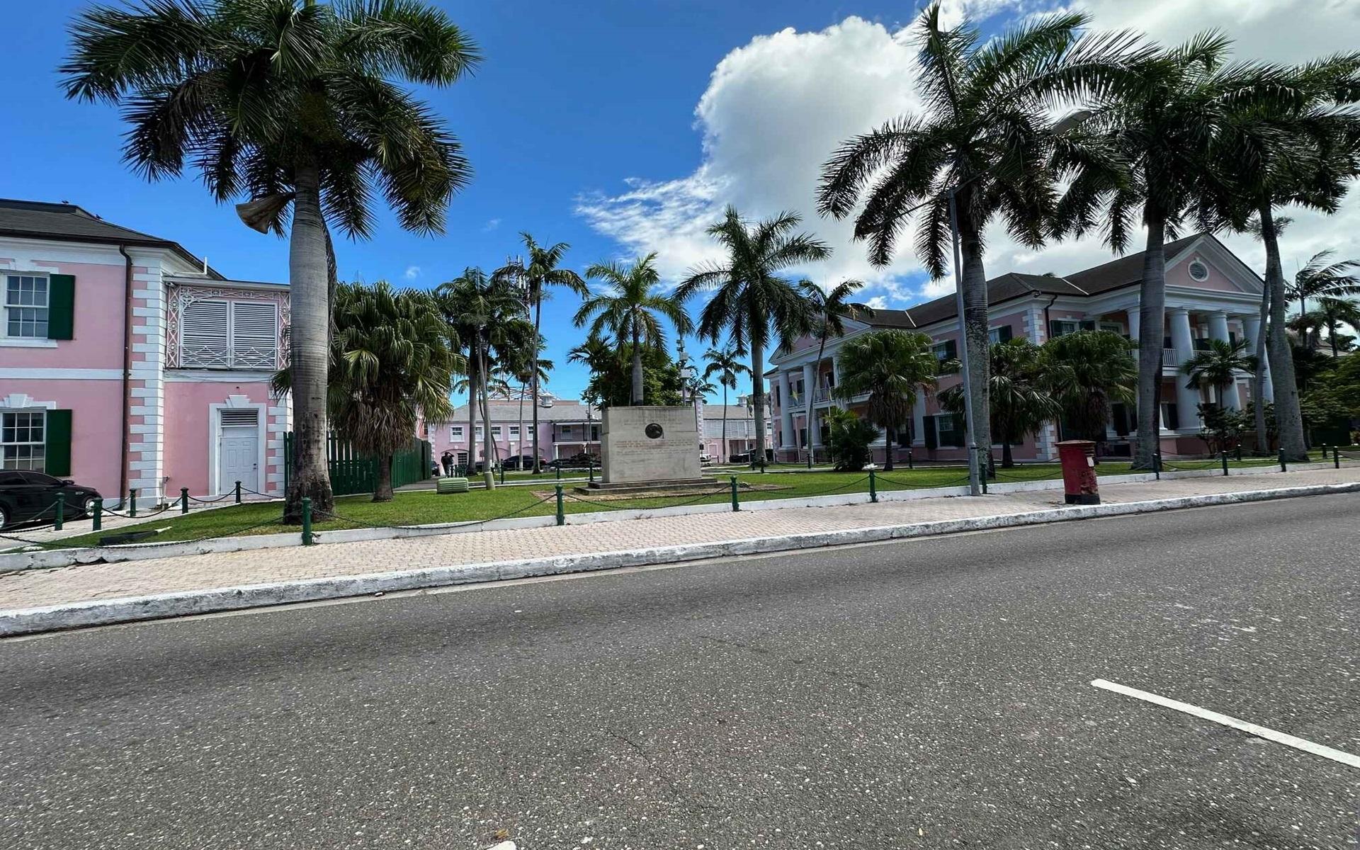 Pink buildings with white columns and green shutters stand among palm trees beside a street.