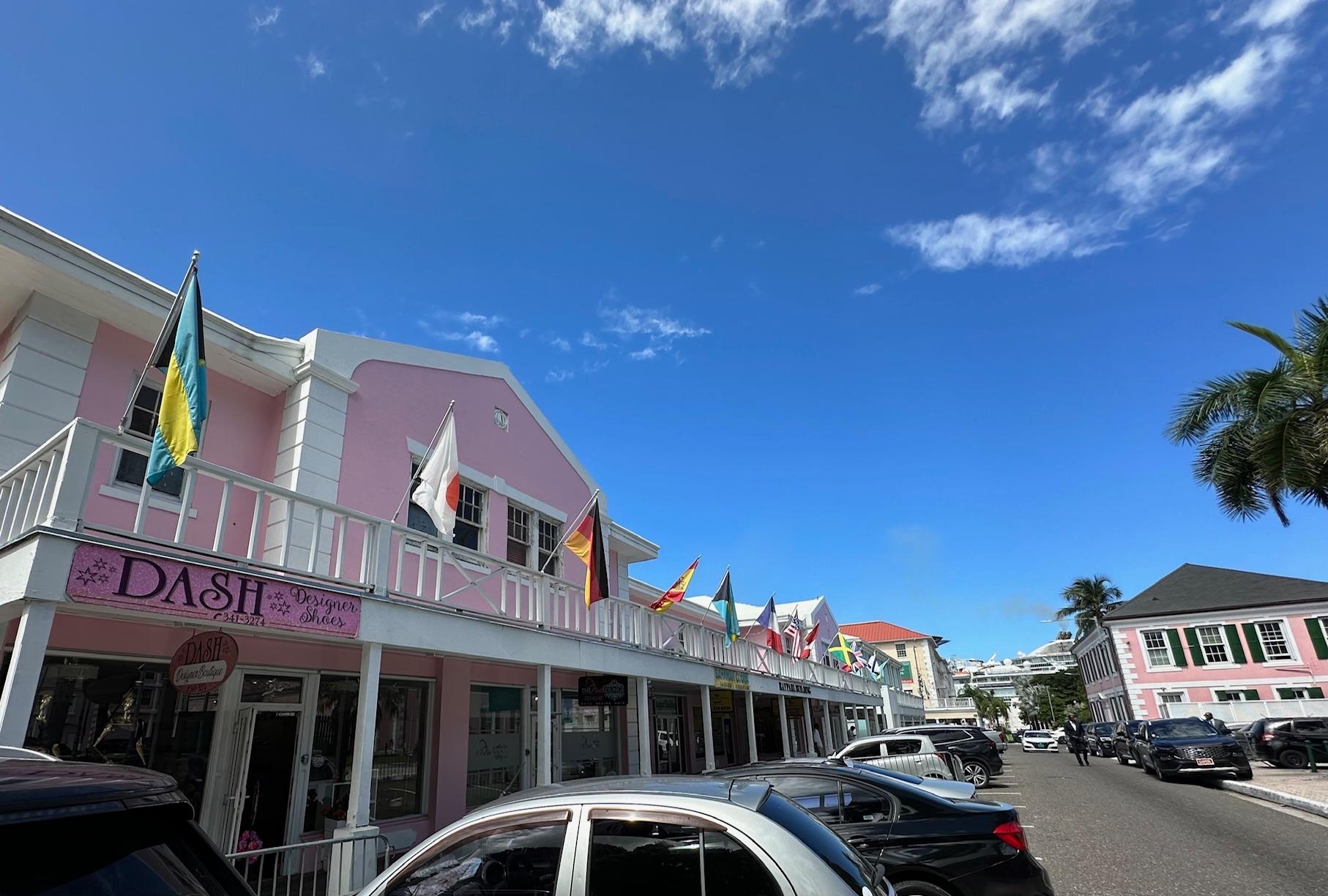 Pink-and-white commercial building with a balcony and flags above street-level shops and parked cars.