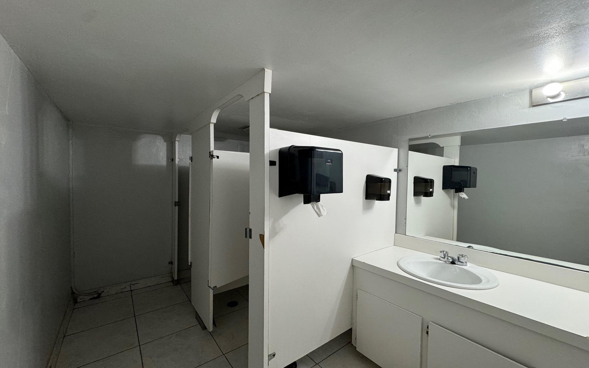 Restroom with white stall partitions, tiled floor, and a sink beneath a large mirror.