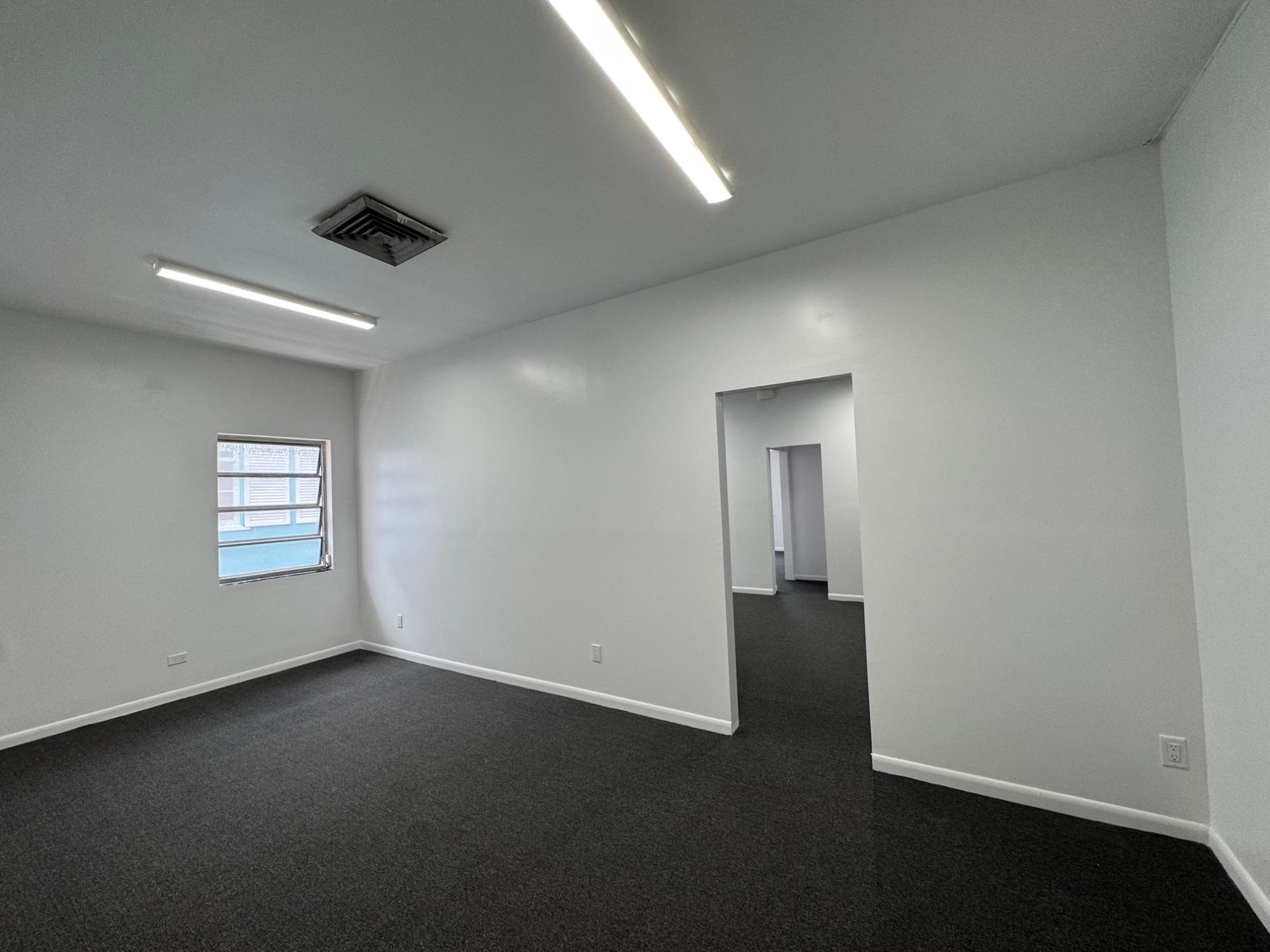 Empty office with dark carpet, fluorescent ceiling lights, a small window, and an open doorway.
