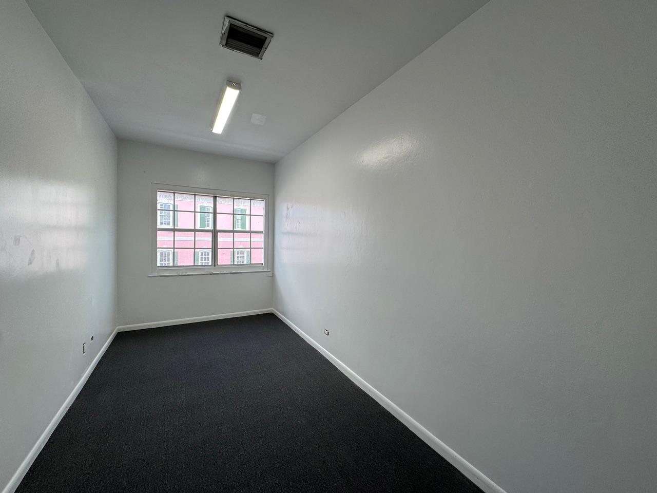 Vacant narrow office room with dark carpet and a window overlooking a pink building.