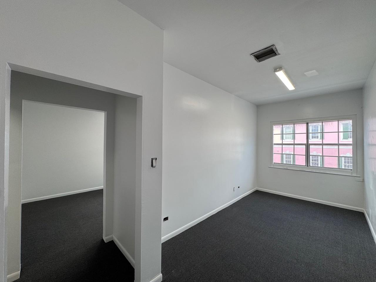 Vacant office with gray carpet, fluorescent ceiling light, and a wide window overlooking a pink building.