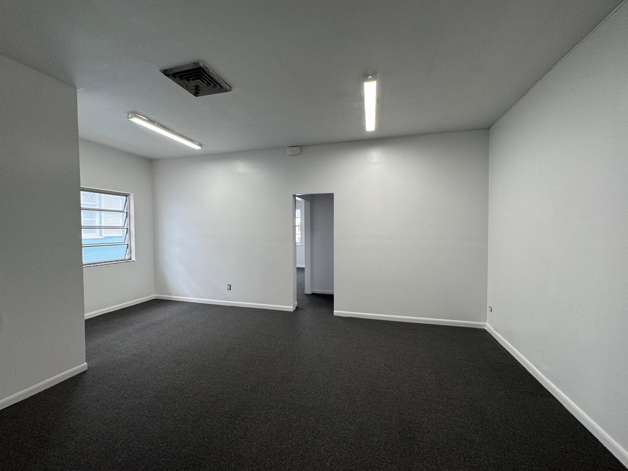 Empty carpeted room with white walls, fluorescent lights, a louvered window, and an adjoining doorway.