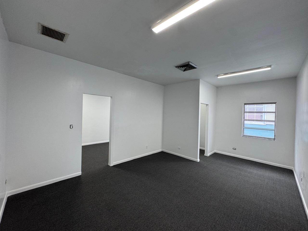 Empty office with dark carpet, fluorescent lights, two doorways, and a small window.