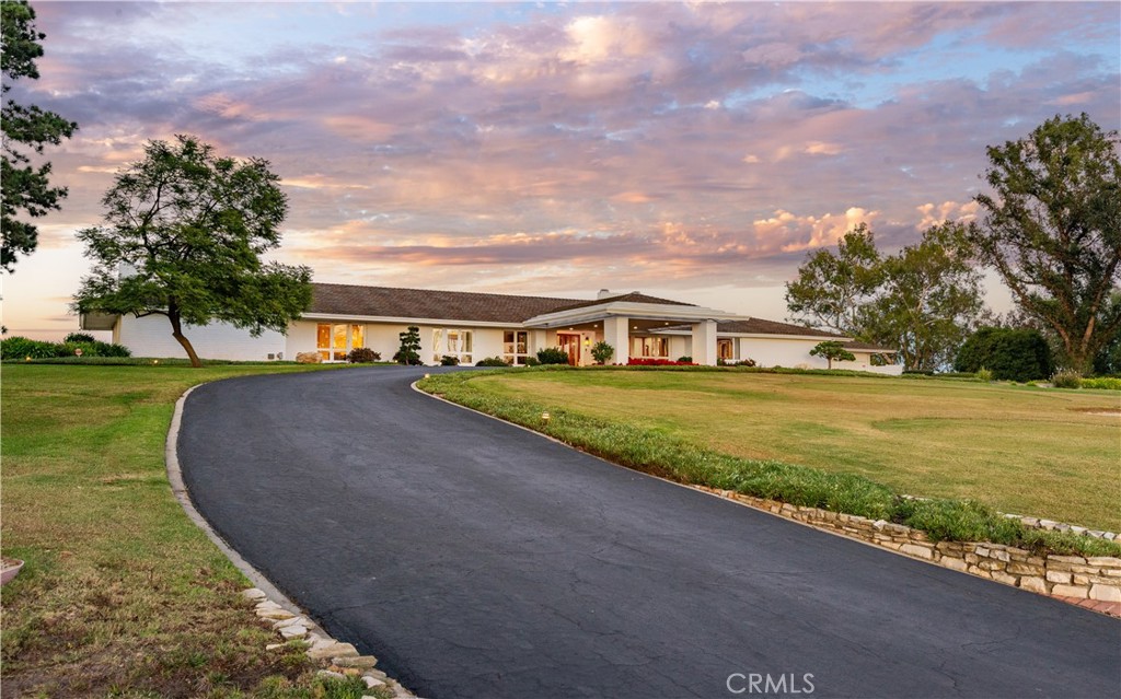 Single-story home behind a curved driveway, with broad lawns and a colorful sunset sky.