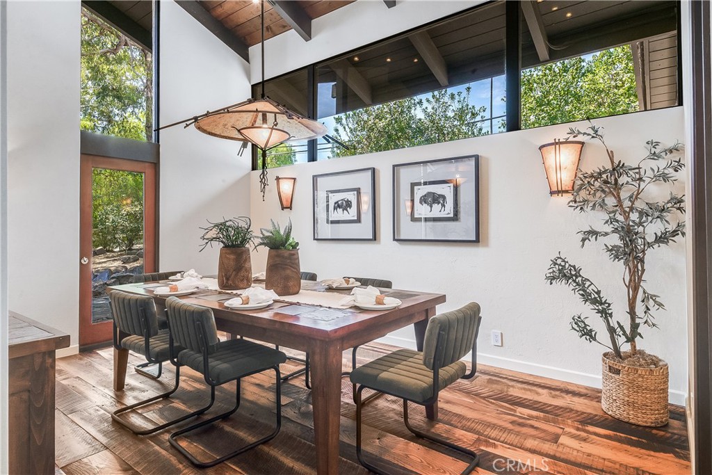 Dining room with a set table, exposed beams, and tall windows overlooking trees.