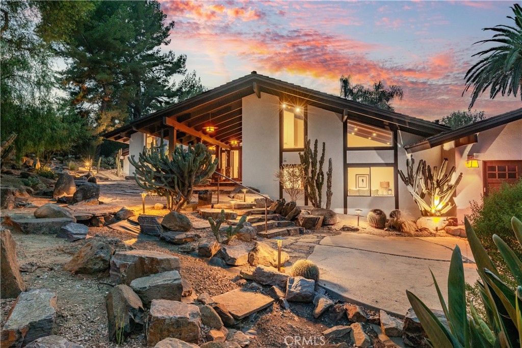 Modern home with angular roof, illuminated windows, and cactus-filled rock landscaping at dusk.