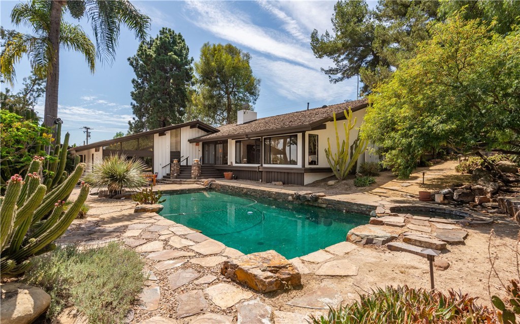 Single-story home behind a turquoise pool, surrounded by stone paving, cacti, and mature trees.