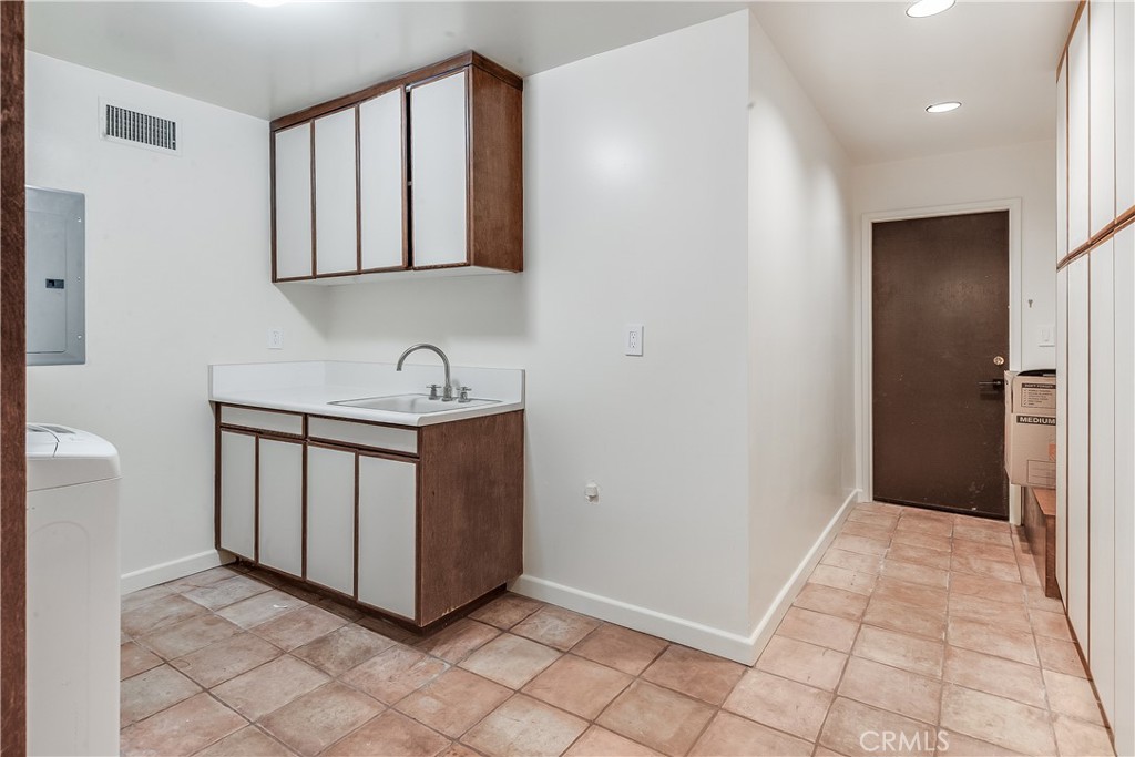 Laundry room with a utility sink, wood-trimmed cabinets, and tiled floor beside a hallway.