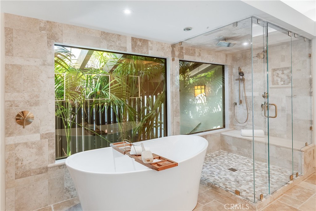 Bathroom with a freestanding soaking tub, glass shower, and windows overlooking tropical greenery.