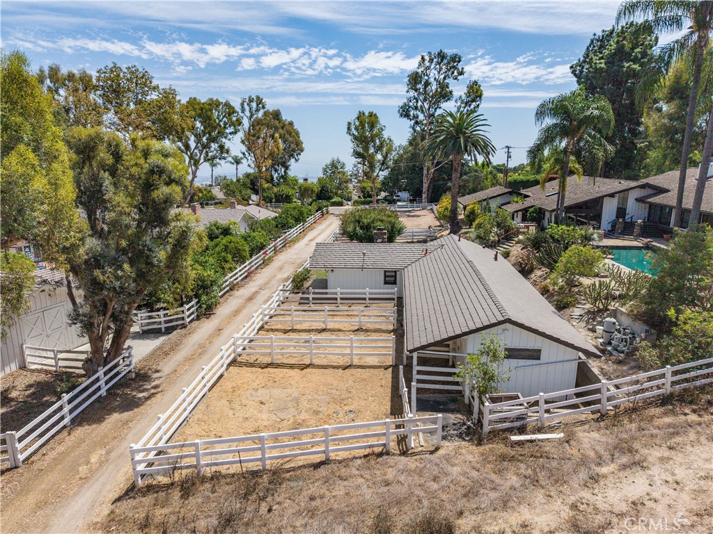 Aerial view of a single-story home, white-fenced paddocks, dirt drive, and neighboring pool.