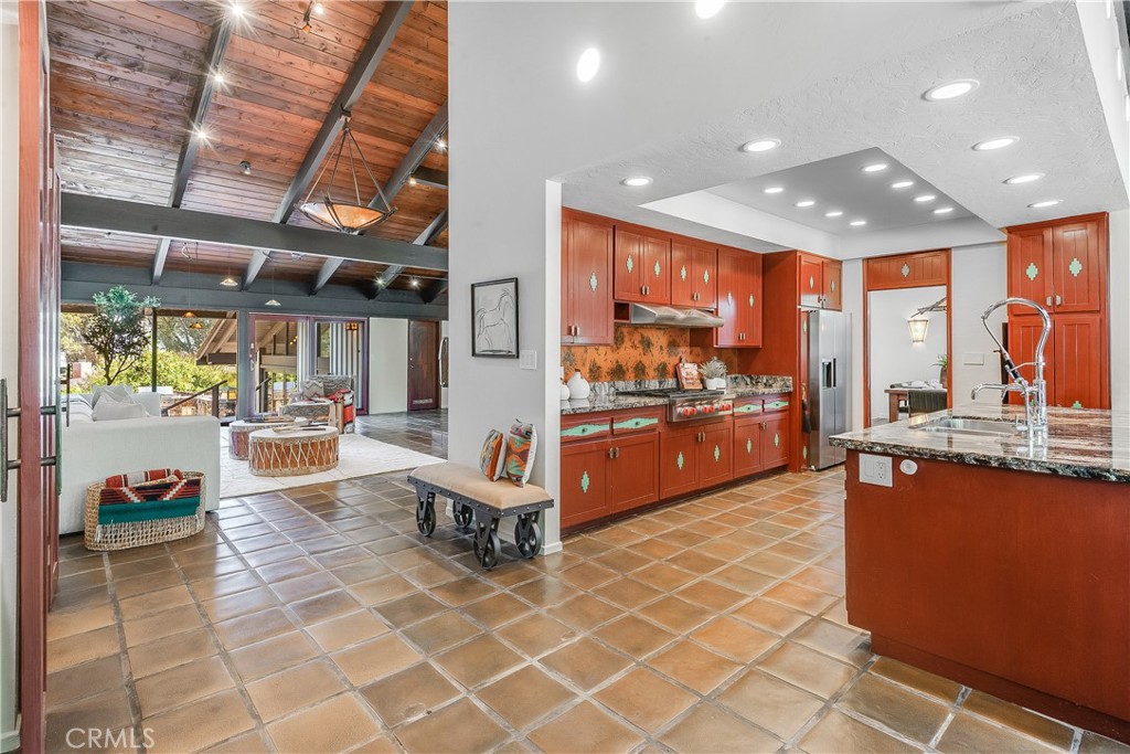 Open kitchen and living room with red wood cabinetry, tiled floors, and an exposed-beam ceiling.