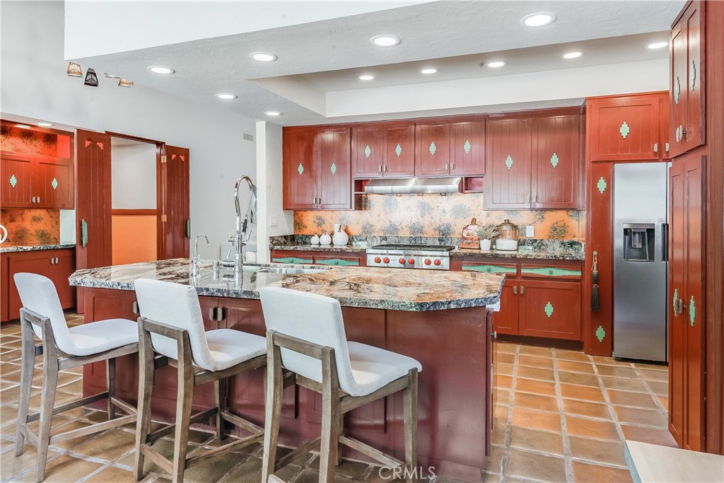 Kitchen with reddish wood cabinetry, a large stone island, stainless steel appliances, and terracotta tile floors.