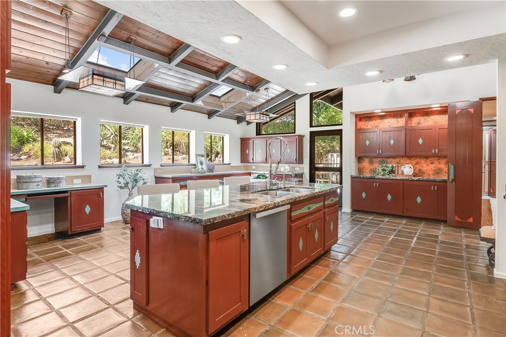 Spacious kitchen with a granite island, reddish wood cabinetry, terracotta tile floors, and an exposed beam ceiling.